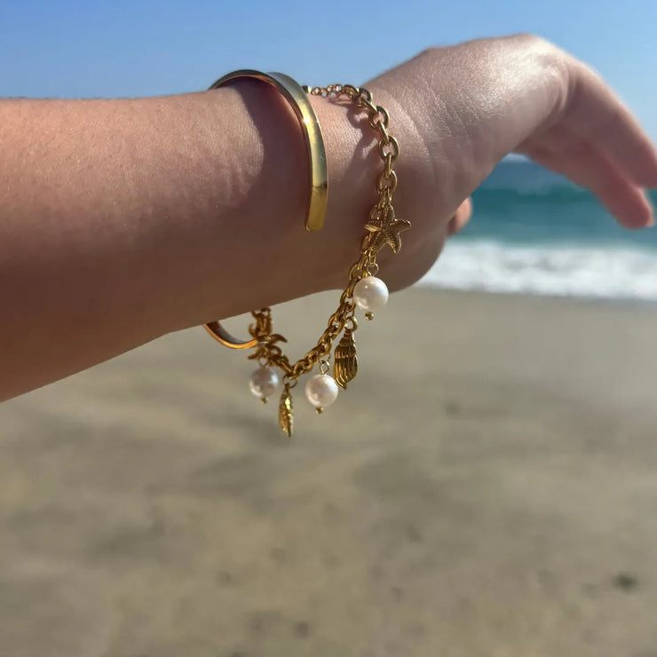 Gold charm bracelet with seashells and pearls, styled for a beach-inspired summer look.