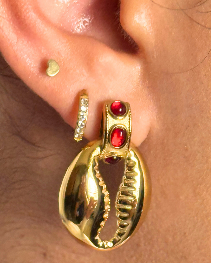 Gold hoop earrings with red gemstones worn on an ear. Red gemstone stud with small seashell charm drop; hypoallergenic, tarnish-free coastal-meets-fall earring