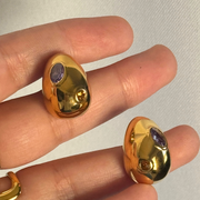 Gold earrings with purple gemstones held on a finger against a plain background. Medium 18k gold-filled oval stud with purple and orange gemstones; hypoallergenic posts; polished fall color pop