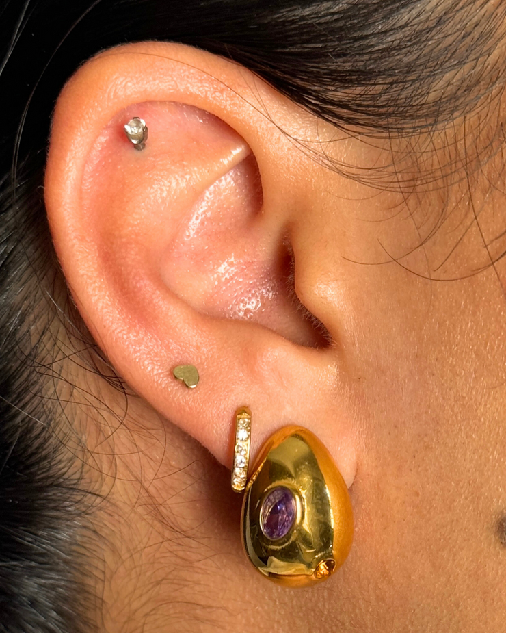 Close-up of an ear wearing a gold earring with a purple gemstone. Medium 18k gold-filled oval stud with purple and orange gemstones; hypoallergenic posts; polished fall color pop