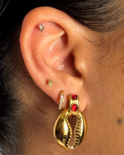 Close-up of an ear with gold earrings featuring red stones.Red gemstone stud with small seashell charm drop; hypoallergenic, tarnish-free coastal-meets-fall earring.