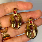 Gold shell-shaped earrings with red stones held in a hand against a neutral background.Red gemstone stud with small seashell charm drop; hypoallergenic, tarnish-free coastal-meets-fall earring